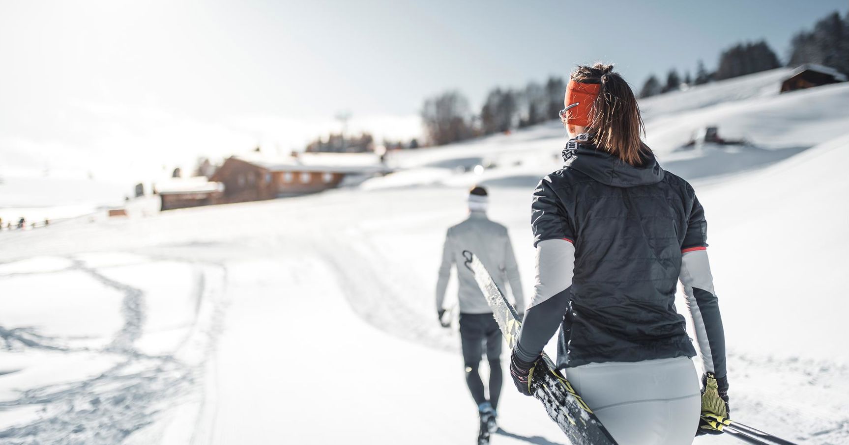 Two cross-country skiers walk through a snowy winter landscape with skis in hand; wooden huts and gentle hills can be seen in the background under a bright blue sky