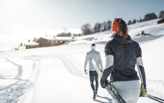 Two cross-country skiers walk through a snowy winter landscape with skis in hand; wooden huts and gentle hills can be seen in the background under a bright blue sky