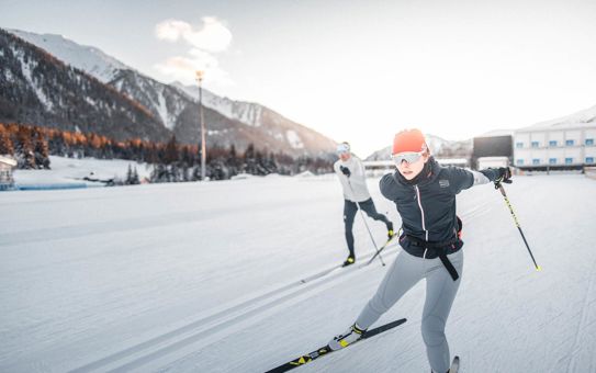 Due fondisti durante l’allenamento nella pista innevata dell’arena del biathlon di Anterselva, in primo piano una donna con fascia rossa, sullo sfondo montagne innevate illuminate dal sole.